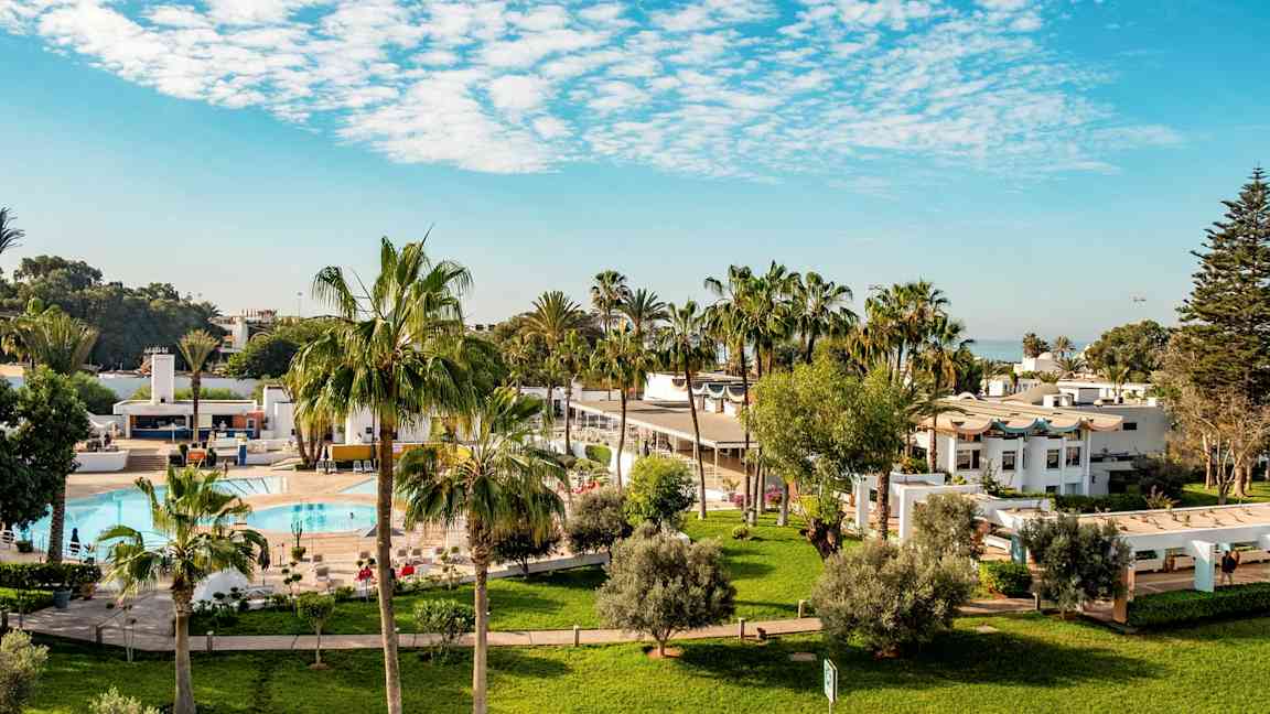 Hotellområde med gräsmattor, palmer, pool och vita byggnader. (AI-genererad text)