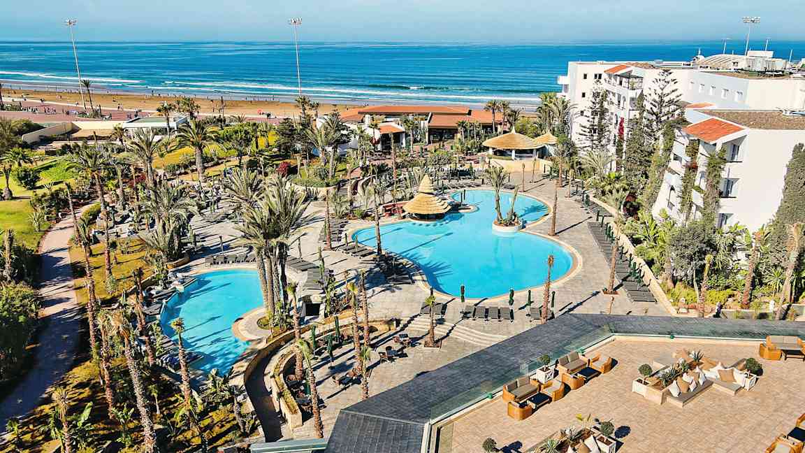 Hotellområde med pooler och omgivande trädgårdar, beläget nära en sandstrand och havet. (AI-genererad text)