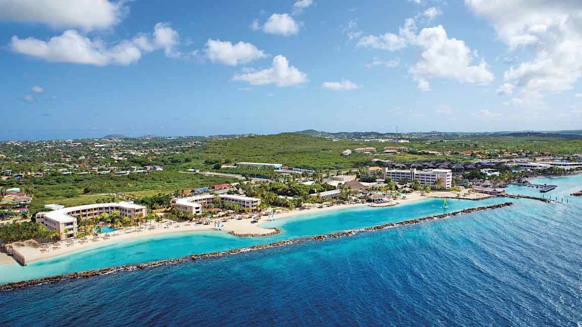 Flygfoto av en kustlinje med hotellbyggnader vid en strand och grönska i bakgrunden. (AI-genererad text)