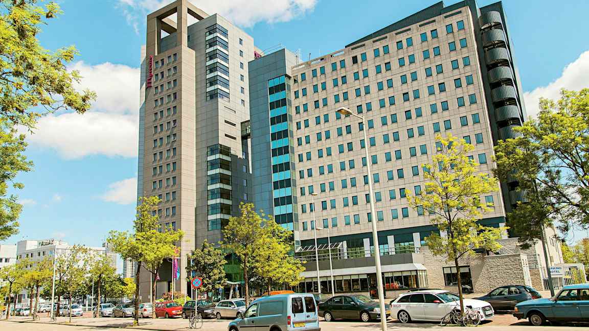 En modern byggnad med flera våningar omgiven av parkerade bilar och träd. (AI-genererad text)