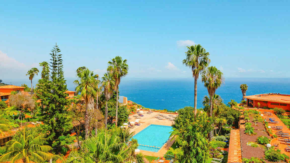 Utsikt över ett poolområde omgivet av grönska och palmer, med havet i bakgrunden. (AI-genererad text)