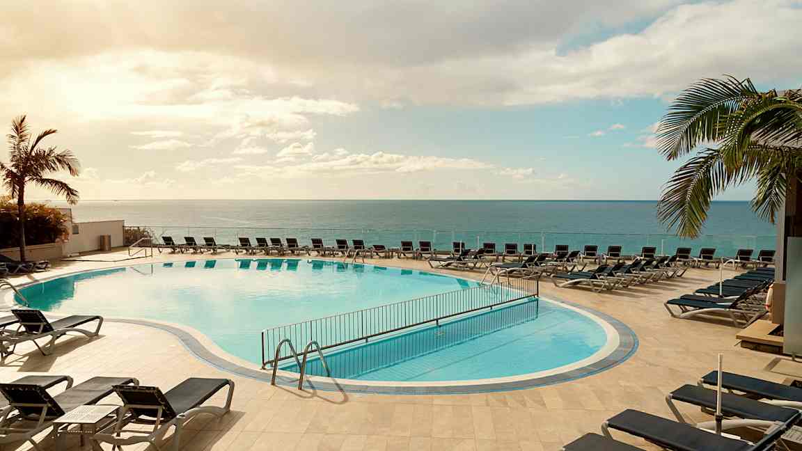 Utomhuspool med solsängar runt omkring, belägen nära havet under en klar himmel. (AI-genererad text)