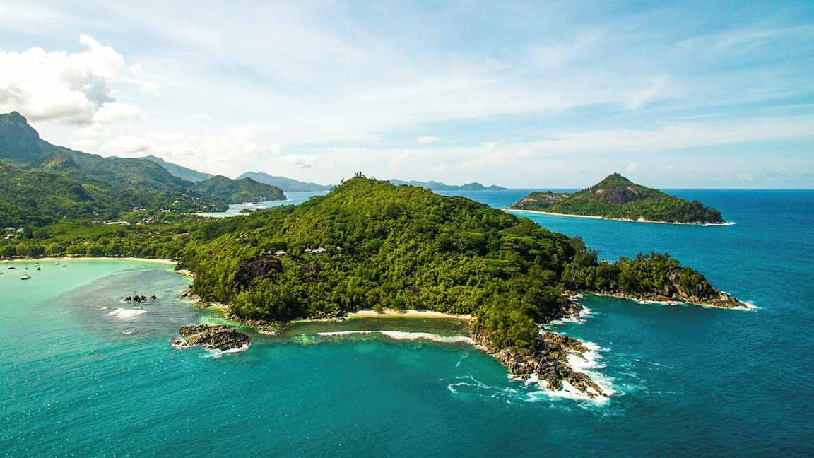 Flygfoto av en grönskande ö omgiven av klarblått hav och mindre öar i bakgrunden. (AI-genererad text)