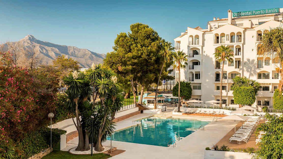 Hotellbyggnad med pool och grönskande omgivning, inramad av växtlighet och ett berg i bakgrunden. (AI-genererad text)