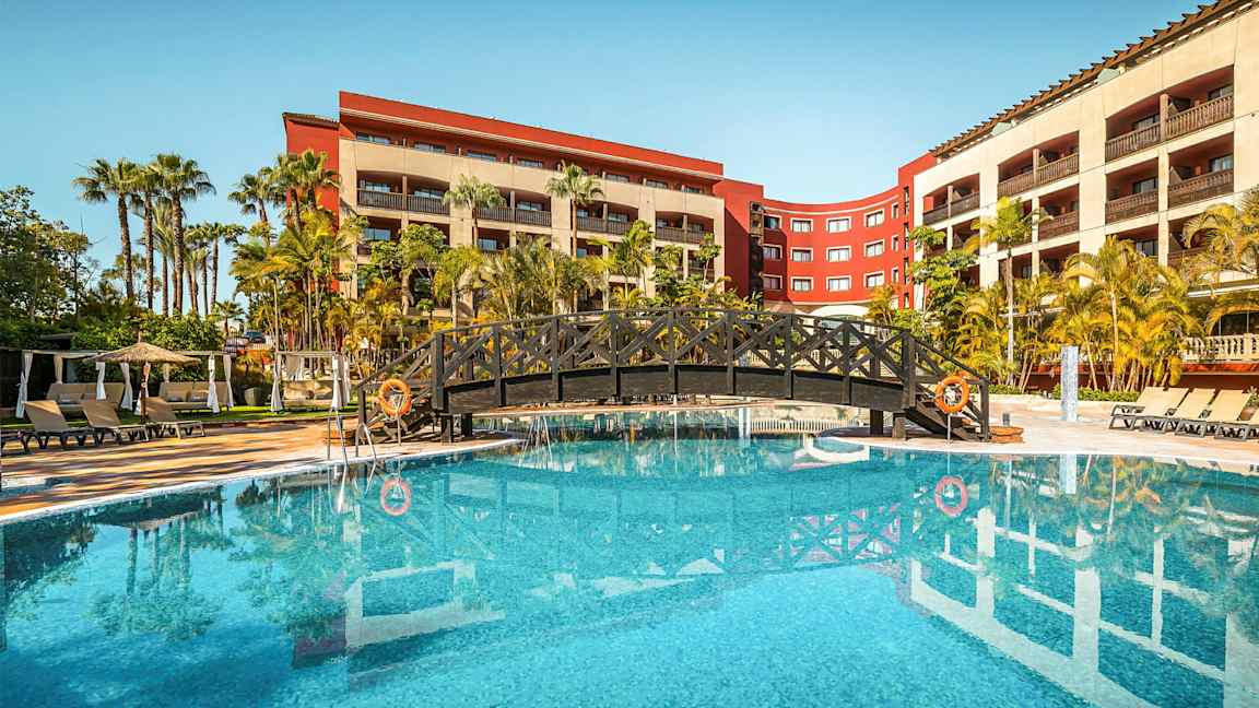 Bild av en hotellpool omgiven av palmer och solstolar med en bro över vattnet. I bakgrunden syns hotellbyggnader. (AI-genererad text)
