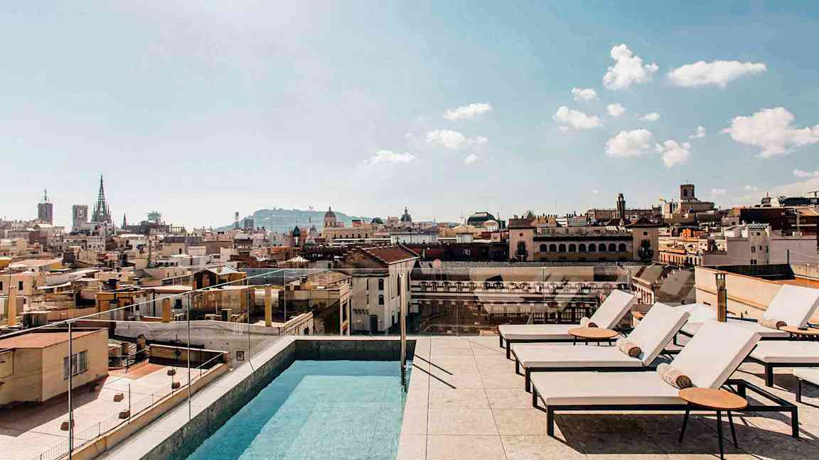 En utomhuspool och solstolar på en takterrass med utsikt över en stad och dess byggnader, under en klar himmel. (AI-genererad text)