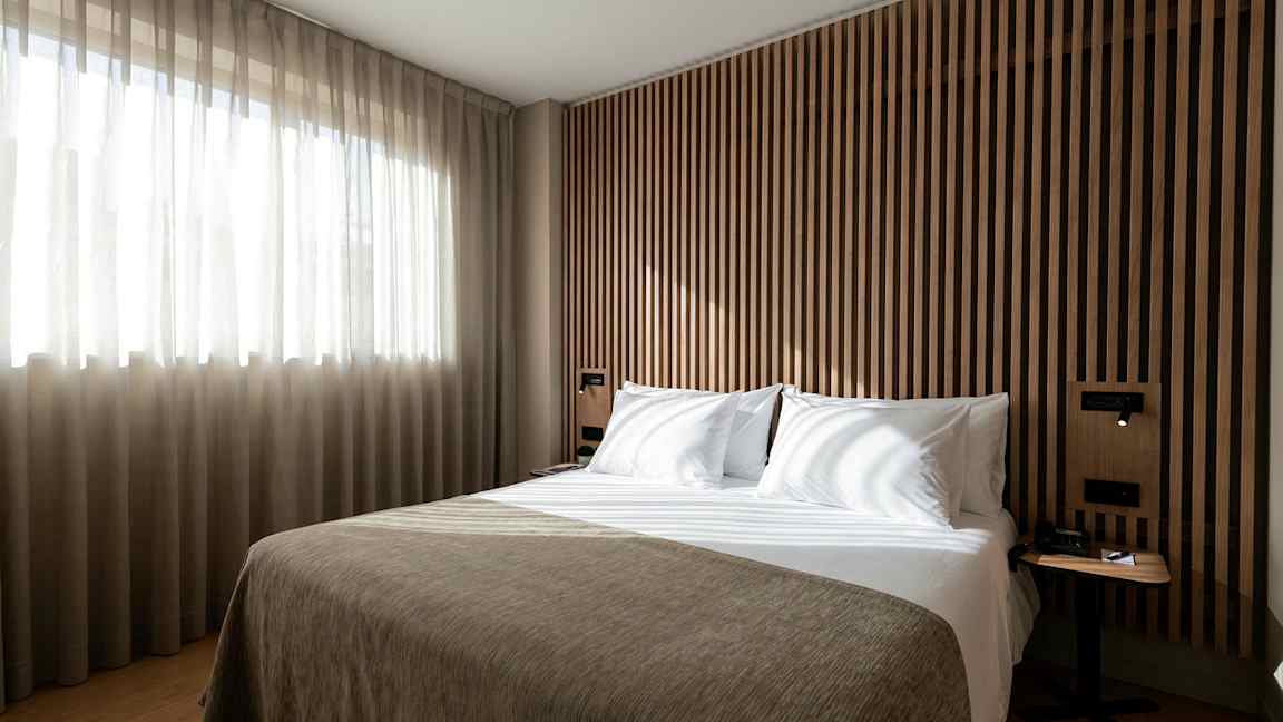 En säng i ett modernt sovrum med träpanel och ljusinsläpp från ett fönster. (AI-genererad text)