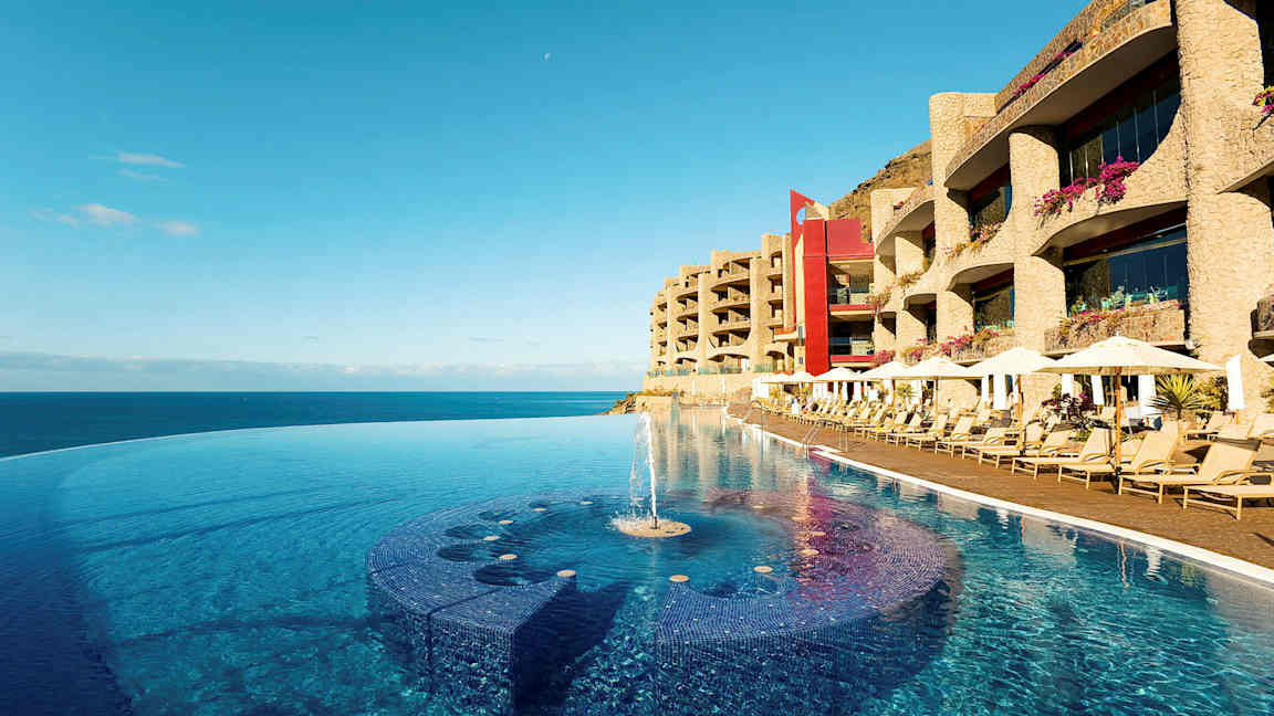 En infinitypool vid strandkanten med solstolar och hotellbyggnader i bakgrunden. (AI-genererad text)