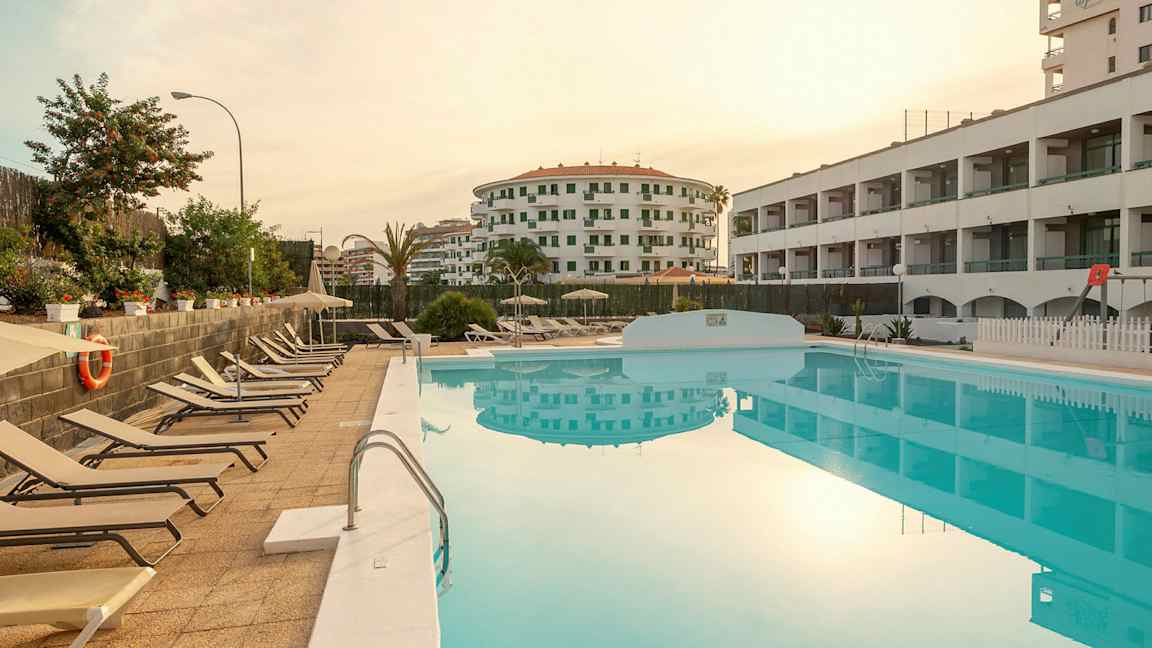 Utsikt över en pool med solstolar bredvid och omgivande byggnader. (AI-genererad text)