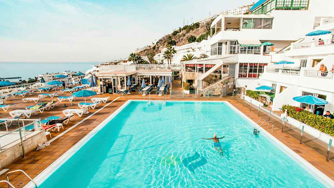 Bild av en stor utomhuspool omgiven av solstolar och parasoller med vita byggnader och hav i bakgrunden. (AI-genererad text)