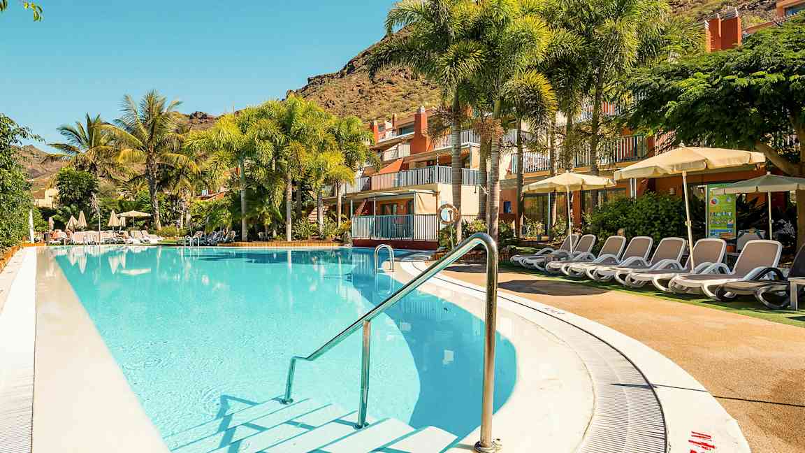 Utomhuspool med solsängar vid ett hotell, omgiven av palmer och gröna växter. (AI-genererad text)