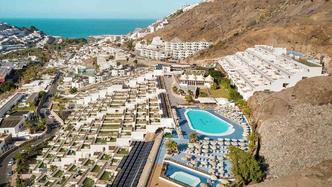Flygfoto av en hotellanläggning med pooler, solstolar och terrasser, belägen vid en sluttning nära kusten. (AI-genererad text)