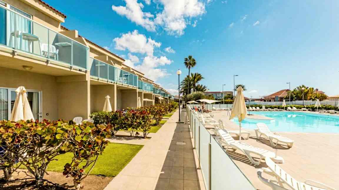 Bild av ett hotellområde med pool, solstolar och grönska. Byggnaden har balkonger. (AI-genererad text)