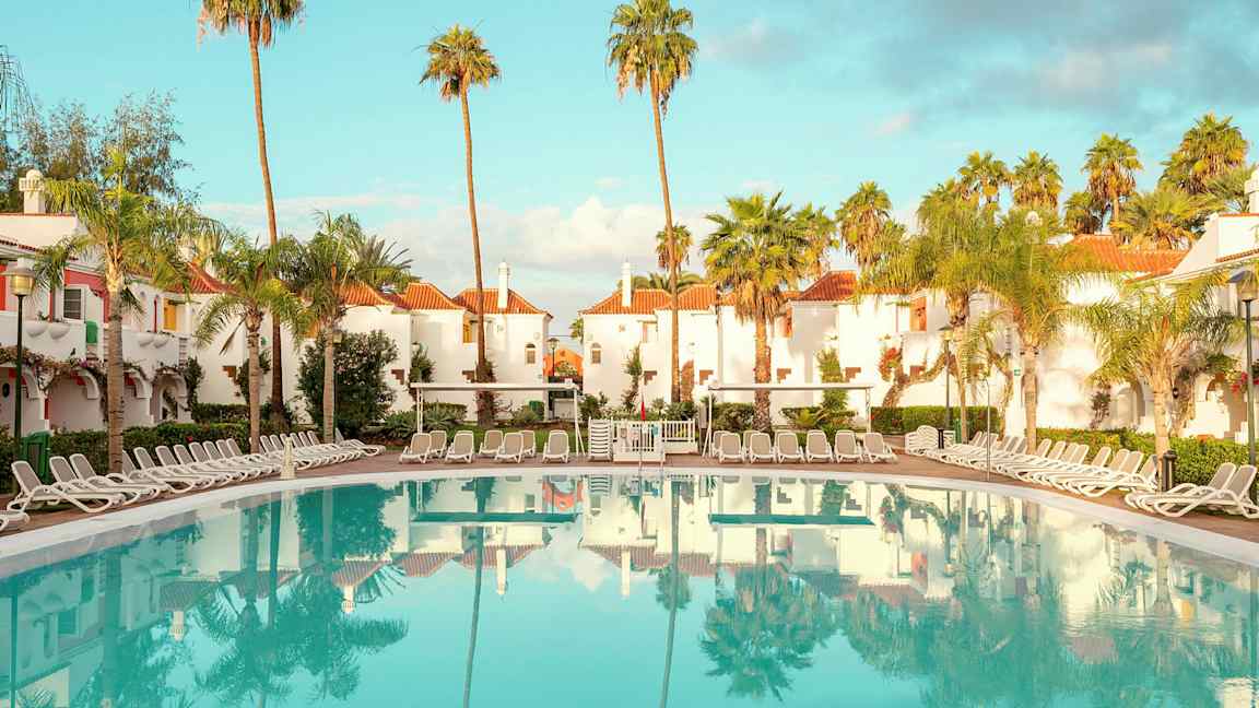 Hotellområde med vitputsade byggnader med röda tak, omgivna av palmer, och en stor pool med solstolar runt. (AI-genererad text)