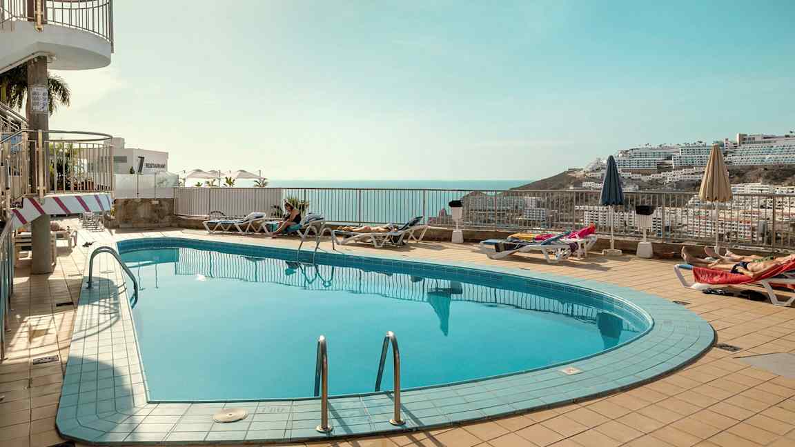 En utomhuspool med solstolar och parasoller runtom, med en vy över havet och omgivande byggnader i bakgrunden. (AI-genererad text)