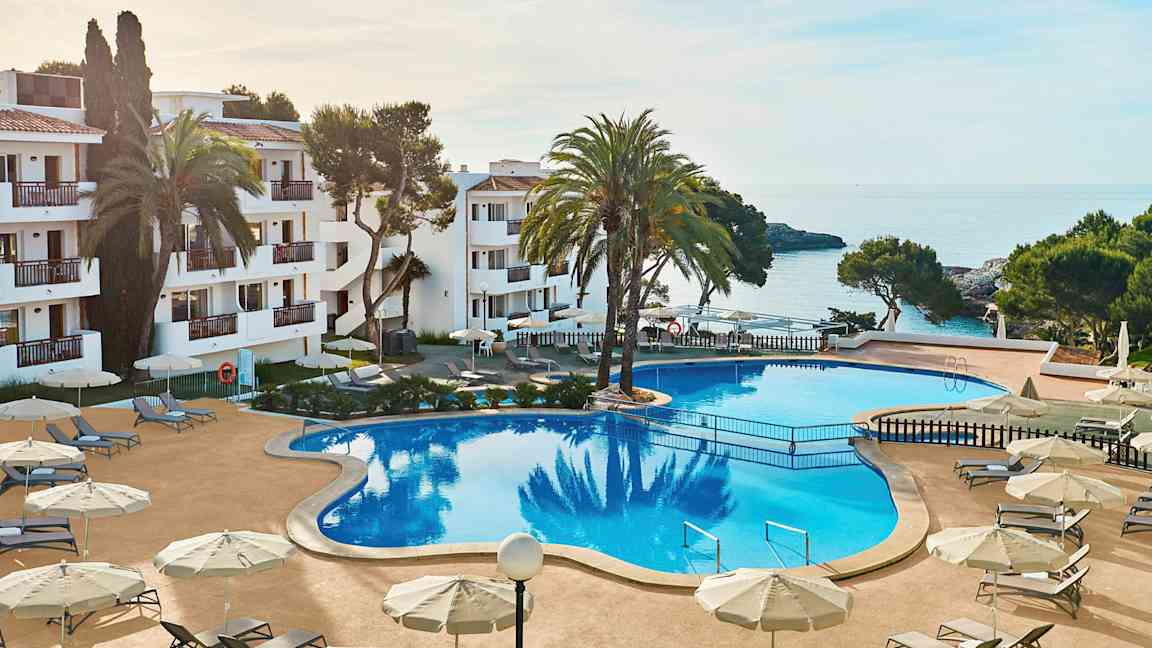 Bild av ett hotellområde med en stor pool, omgiven av solstolar och parasoller. I bakgrunden syns havet och delar av byggnaden. (AI-genererad text)