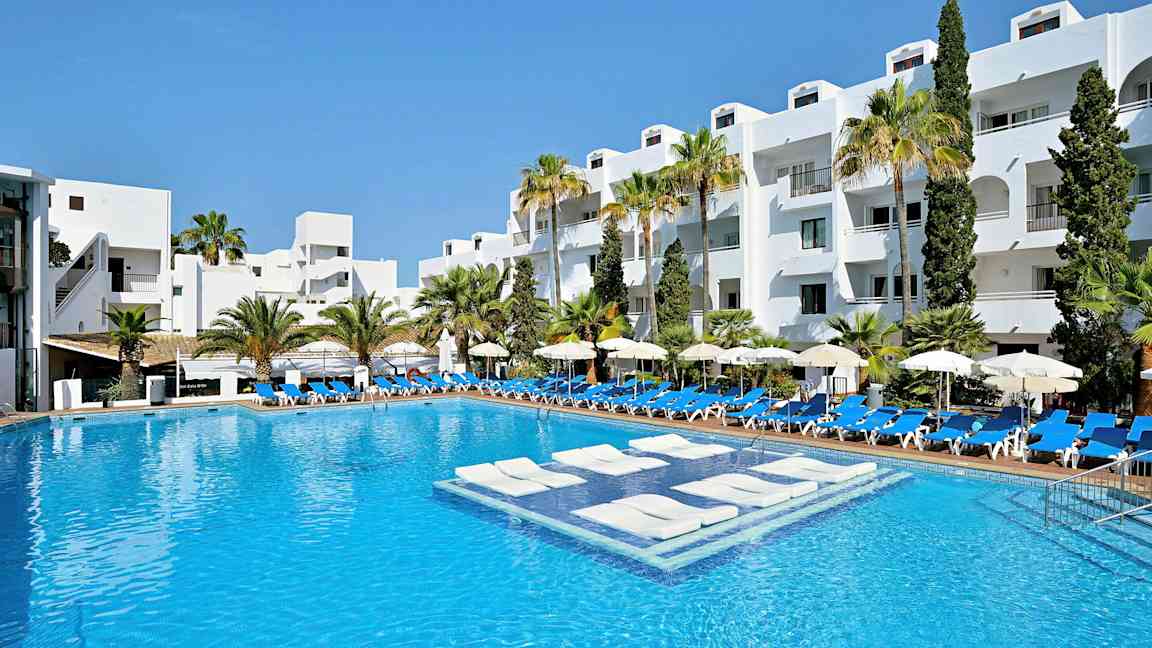 En hotellpool med vitmålade byggnader och palmer i bakgrunden. (AI-genererad text)