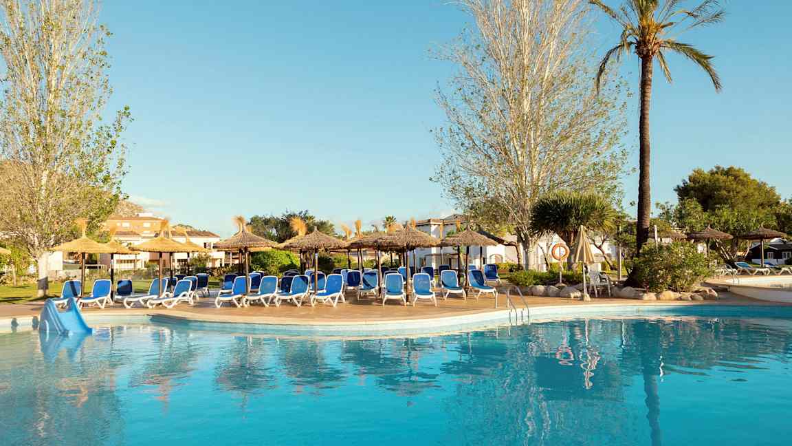 En pool vid ett hotell omgiven av solstolar och parasoller, omgärdad av träd och grönska. (AI-genererad text)