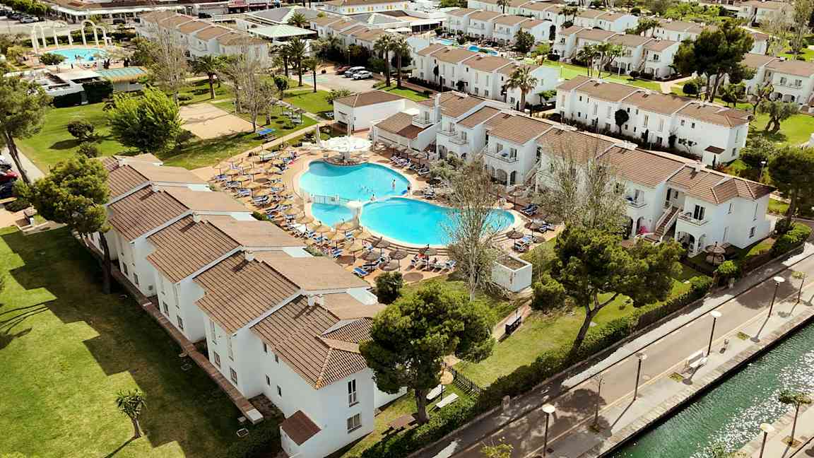 En flygbild av ett semesterkomplex med vita byggnader, en pool och grönområden. (AI-genererad text)