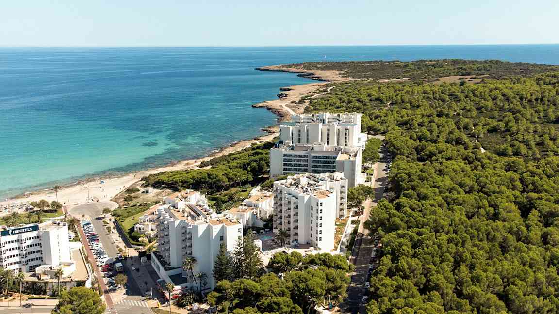 Flygfoto av en kustlinje med hotellbyggnader framför en skog och nära en strand. (AI-genererad text)