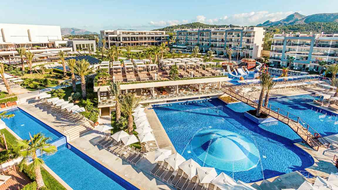 En vy över ett hotellområde med pooler, solstolar och byggnader. (AI-genererad text)