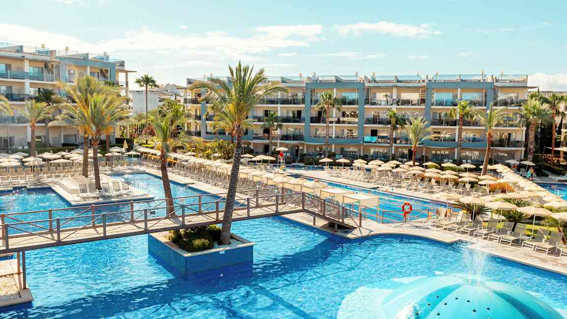 Poolområde vid ett hotell med solstolar och parasoller, omgivet av palmer och byggnader. (AI-genererad text)