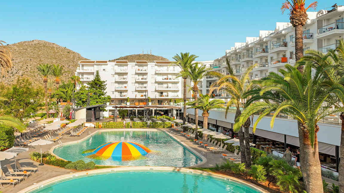 Ett hotell med vita byggnader, solstolar och en pool med en färgglad vattensprutande kupol i mitten. (AI-genererad text)