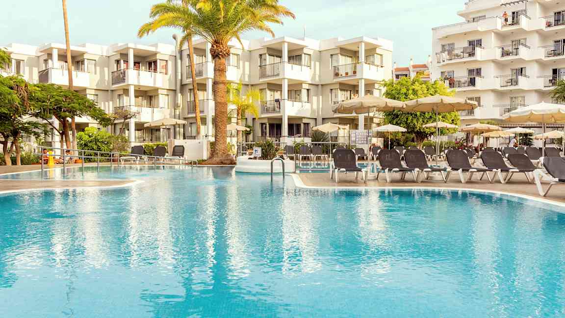 Utomhuspool omgiven av solstolar och parasoller, med en modern byggnad i bakgrunden. (AI-genererad text)