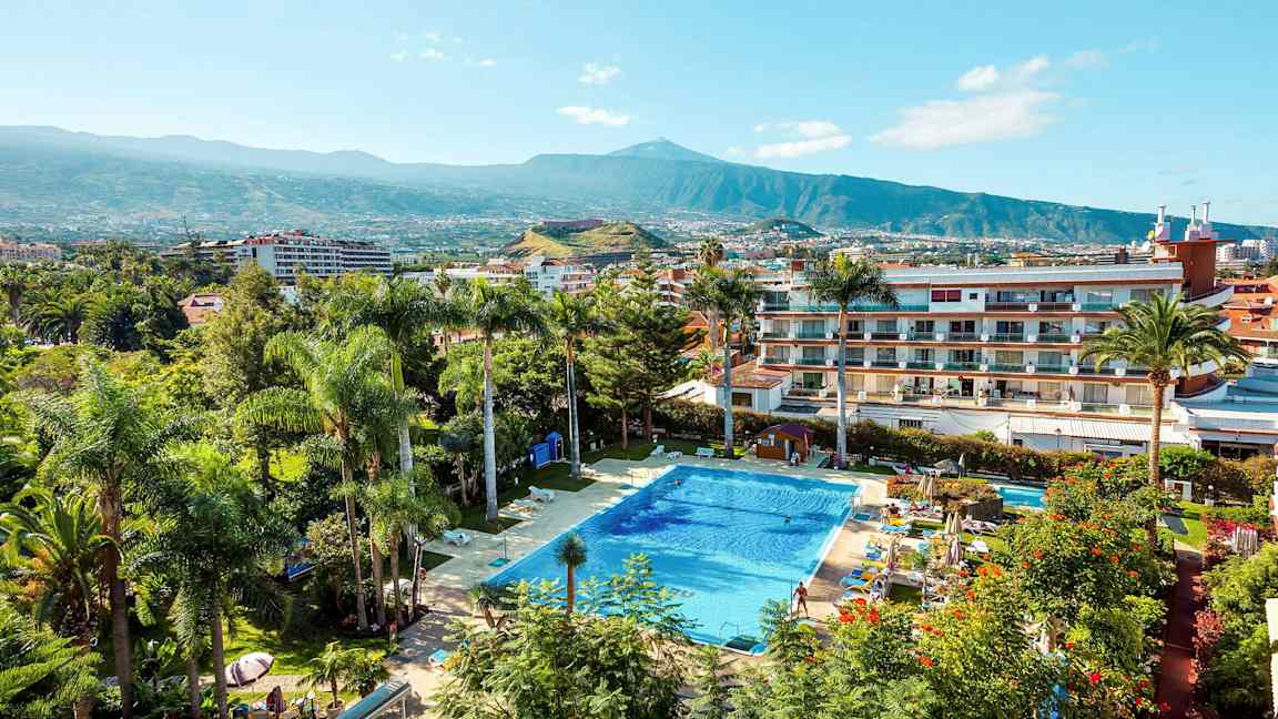 En pool omgiven av växter och byggnader med ett berg i bakgrunden. (AI-genererad text)