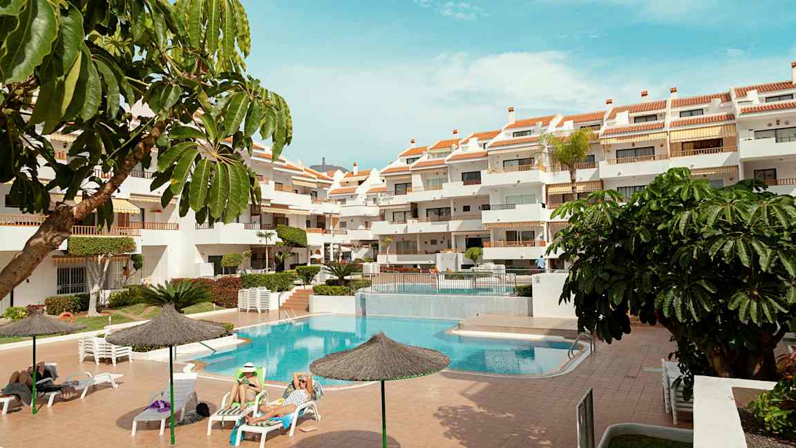 Ett utomhusområde med en pool, solstolar och ett flervåningshotell. Flera parasoller och gröna växter omger området. (AI-genererad text)