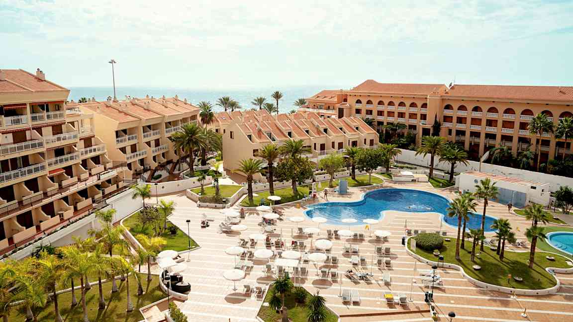 Flera byggnader med terrakottatak omger en pool och solstolar, med omgivande palmer och havet i bakgrunden. (AI-genererad text)