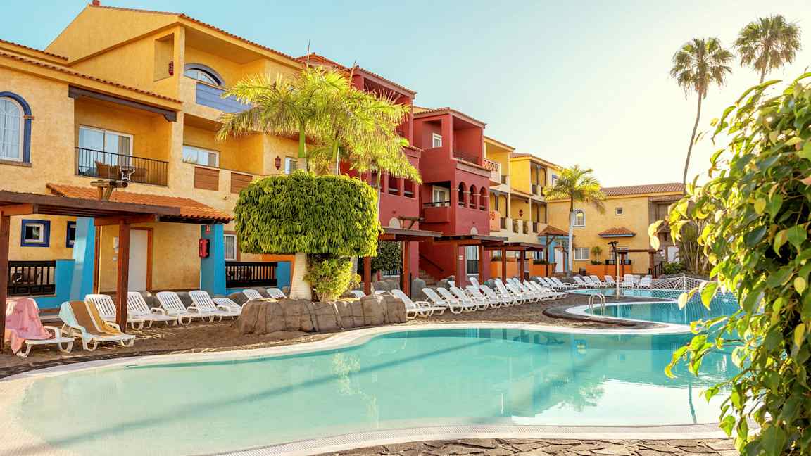 Poolområde med solstolar framför färgglada byggnader. (AI-genererad text)