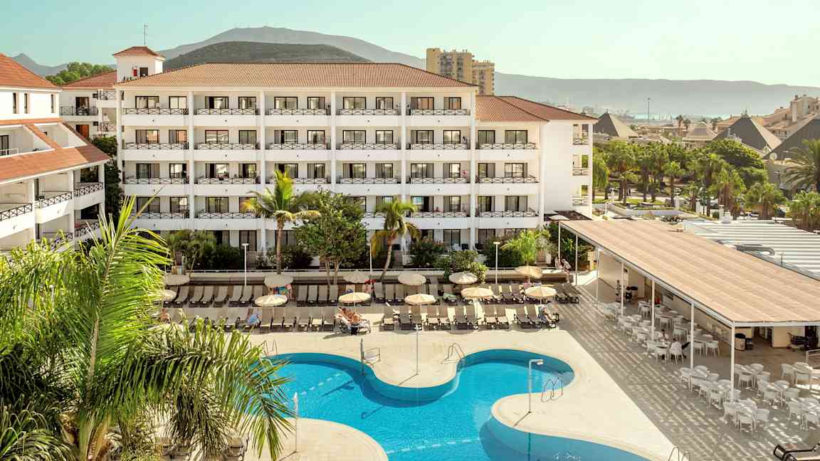 Hotellkomplex med bassäng och solstolar, omgiven av grönska och berg i bakgrunden. (AI-genererad text)