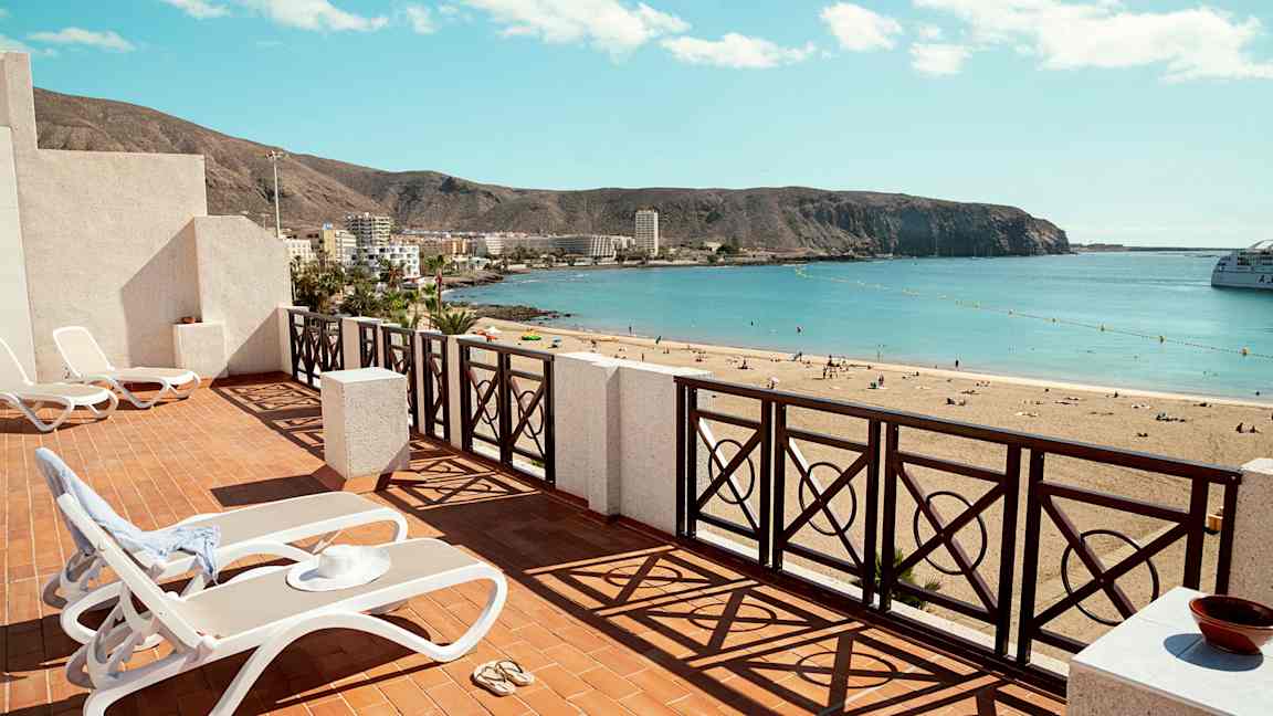 Terrass med solstolar och utsikt över en sandstrand med havet vid horisonten. (AI-genererad text)