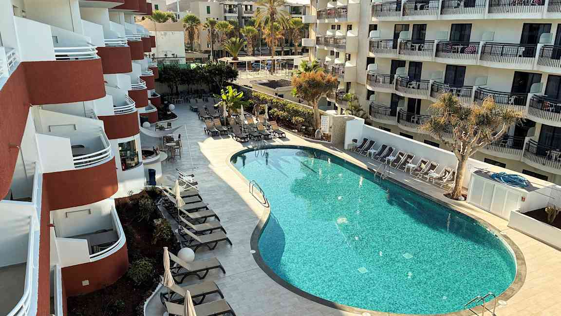 Ett hotell med flera balkonger och en formad pool omgiven av solstolar och parasoller. (AI-genererad text)