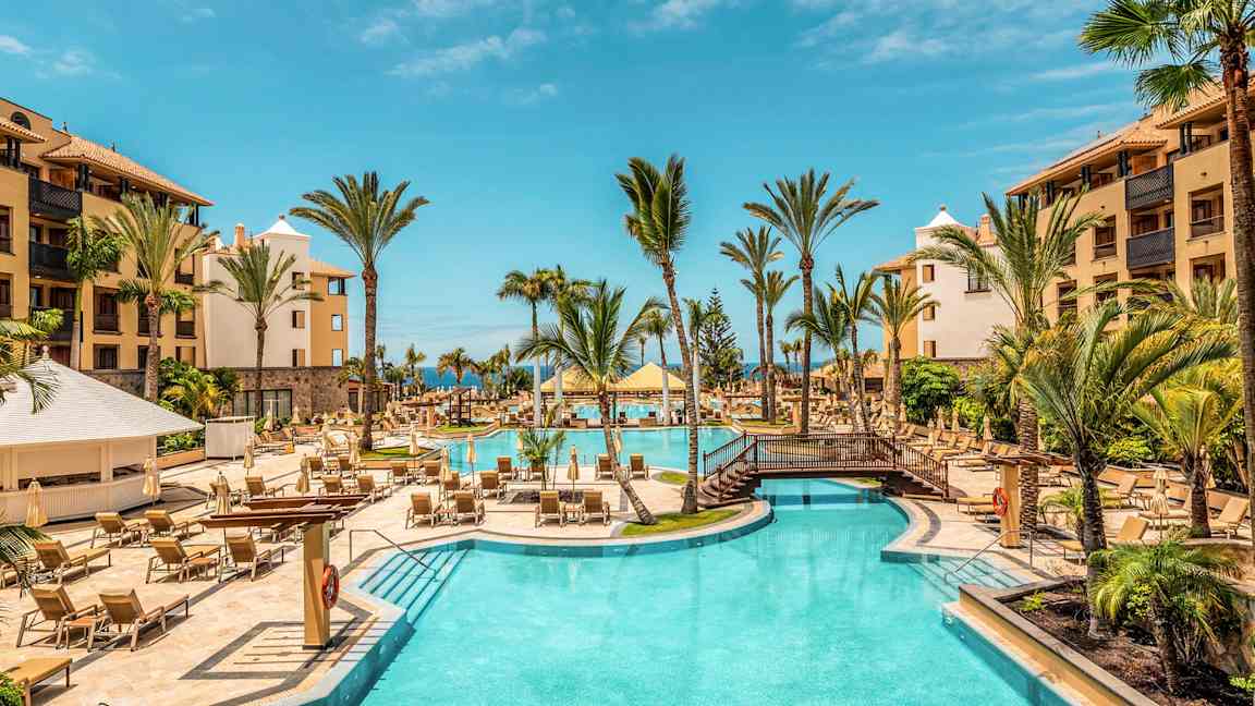 Hotellpool utomhus med palmer och solstolar, omgiven av byggnader. (AI-genererad text)