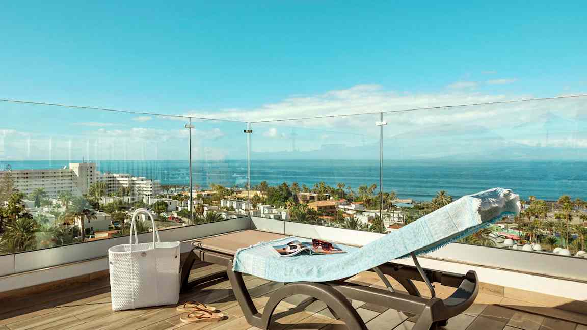 Utsikt från en terrass med en solstol klädd med en handduk, en vit väska och sandaler, över ser man havet och bebyggelse. (AI-genererad text)