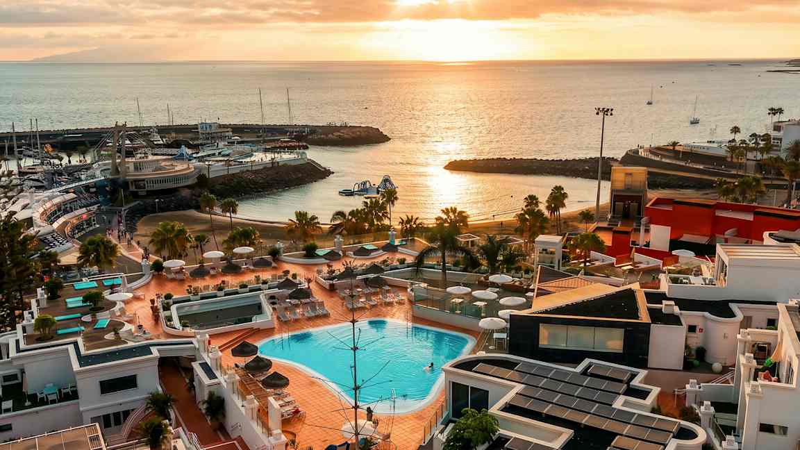 En vy över en resort med pooler, solstolar och paraplyer, med en strand och småbåtshamn vid kusten. Solnedgångens ljus reflekteras i vattnet. (AI-genererad text)