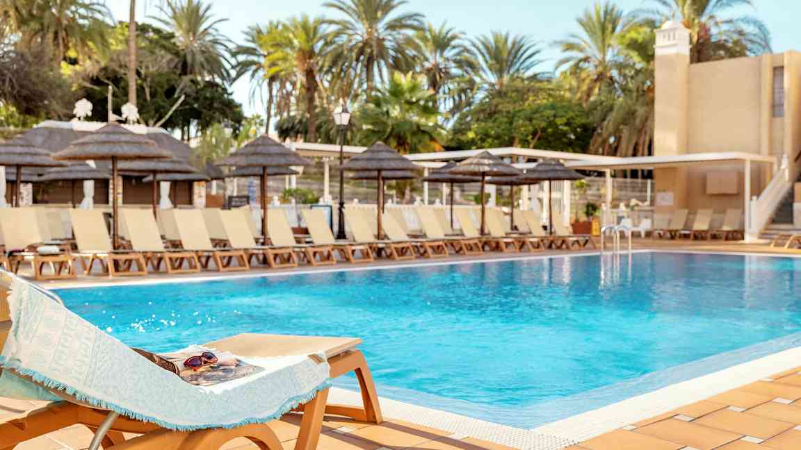 En pool omgiven av solstolar och palmer under en klar himmel. (AI-genererad text)