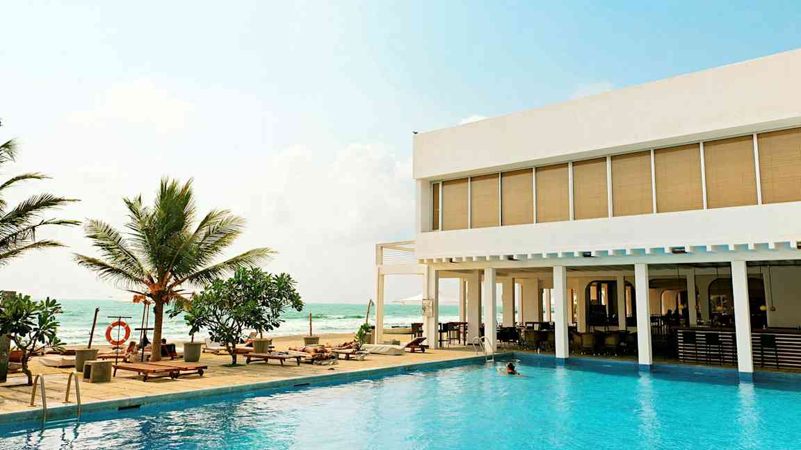 En utomhuspool med solstolar och palmer vid sidan av ett modernt hotell med utsikt över havet. (AI-genererad text)