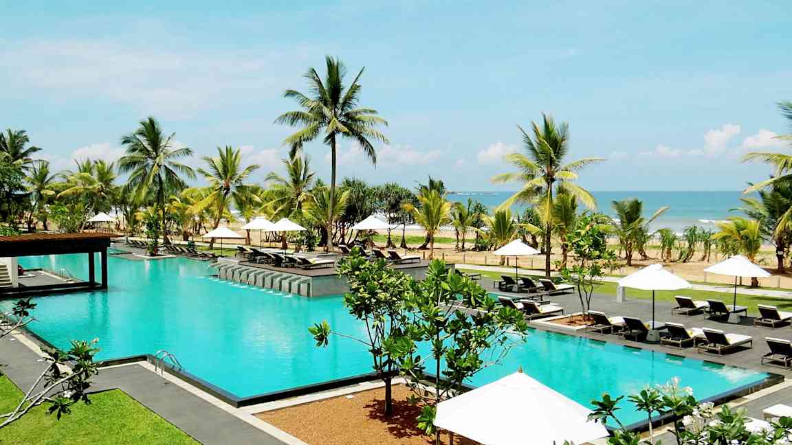 En pool utomhus omgiven av solstolar, parasoller, palmer och med utsikt över havet. (AI-genererad text)