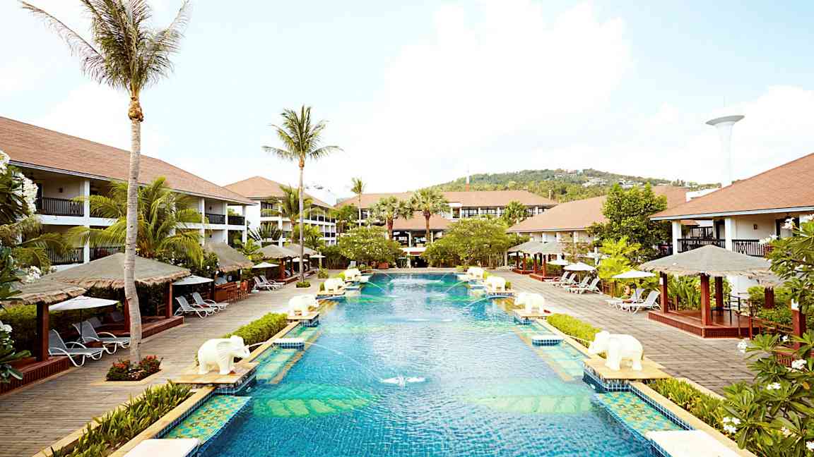 En stor pool på ett semesterortsområde omgiven av palmer, solstolar och bungalowliknande byggnader. (AI-genererad text)