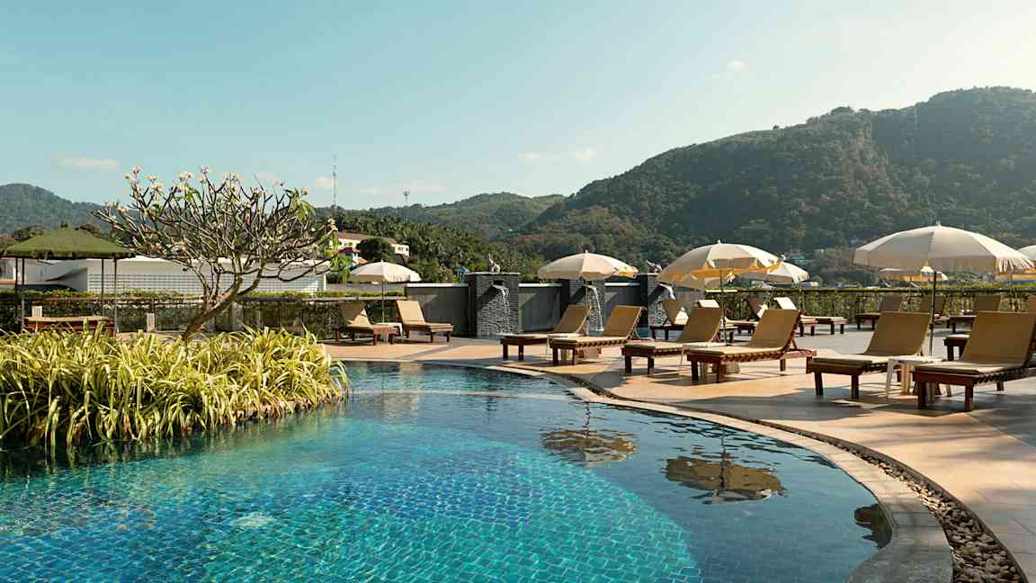 Utomhuspool med solstolar och parasoller, omgiven av grönska och berg i bakgrunden. (AI-genererad text)