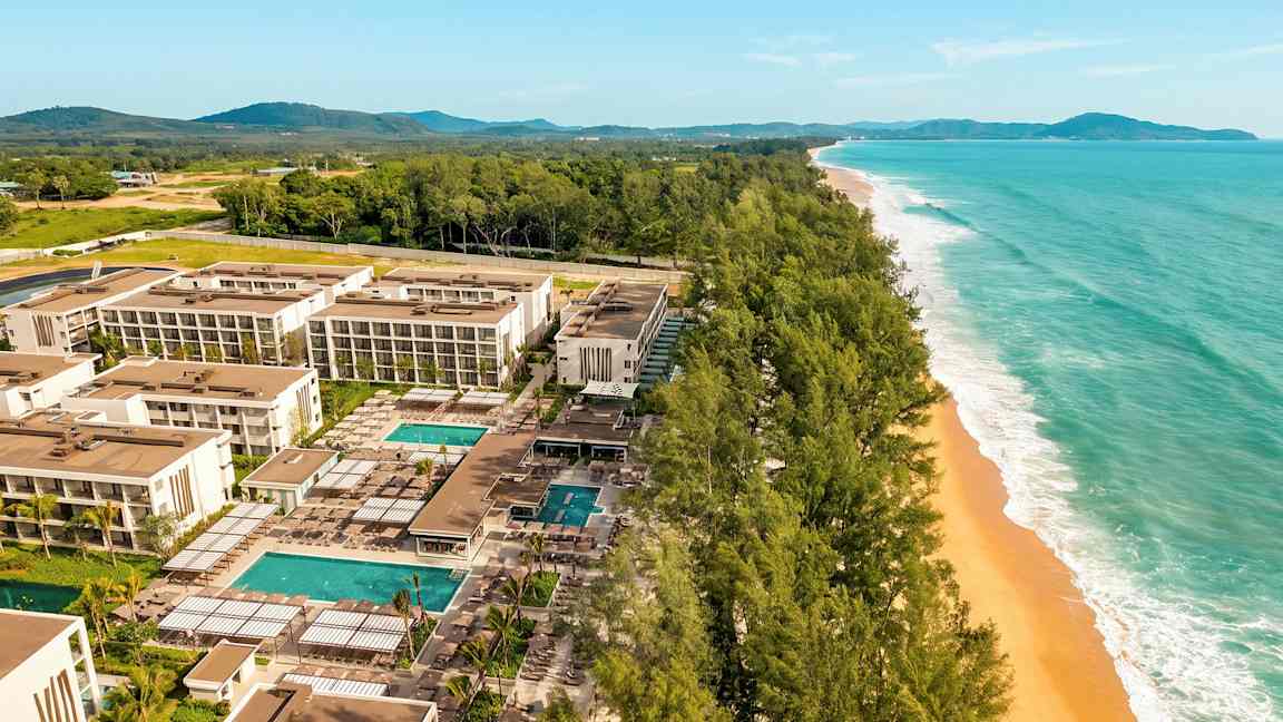 Flygfoto över ett modernt hotellkomplex med flera byggnader, pooler och trädgårdar, beläget vid en bred strand med gyllene sand och omgiven av gröna träd. (AI-genererad text)