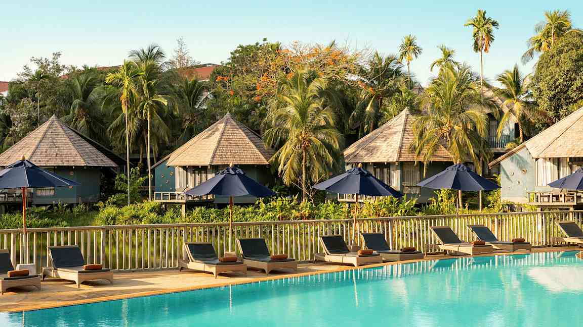 Solstolar och parasoller vid en swimmingpool med bungalowliknande byggnader och grönska i bakgrunden. (AI-genererad text)