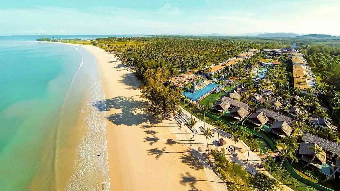 Flygfoto av en lång sandstrand med turkost vatten och en semesteranläggning med bungalower och pooler. (AI-genererad text)