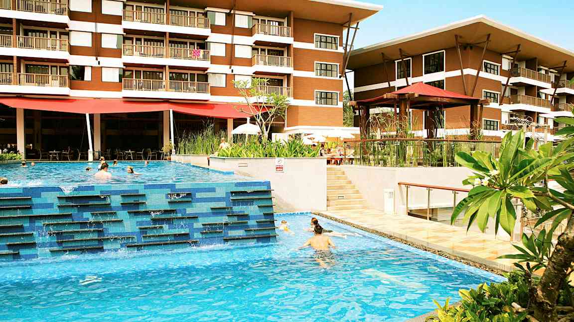 Ett hotell med två våningar syns bakom en utomhuspool där människor simmar. Gröna växter och terrassområden omger poolområdet. (AI-genererad text)