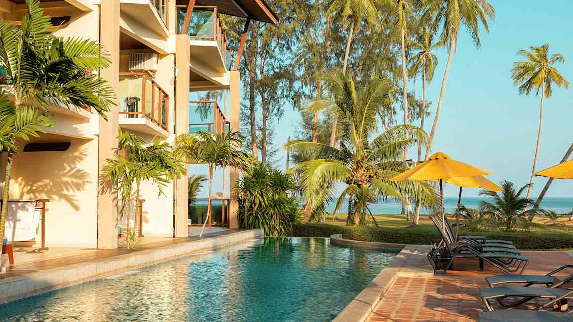 En utomhuspool bredvid ett hotell med balkonger, omgiven av tropiska palmer och gräsyta vid havet. (AI-genererad text)