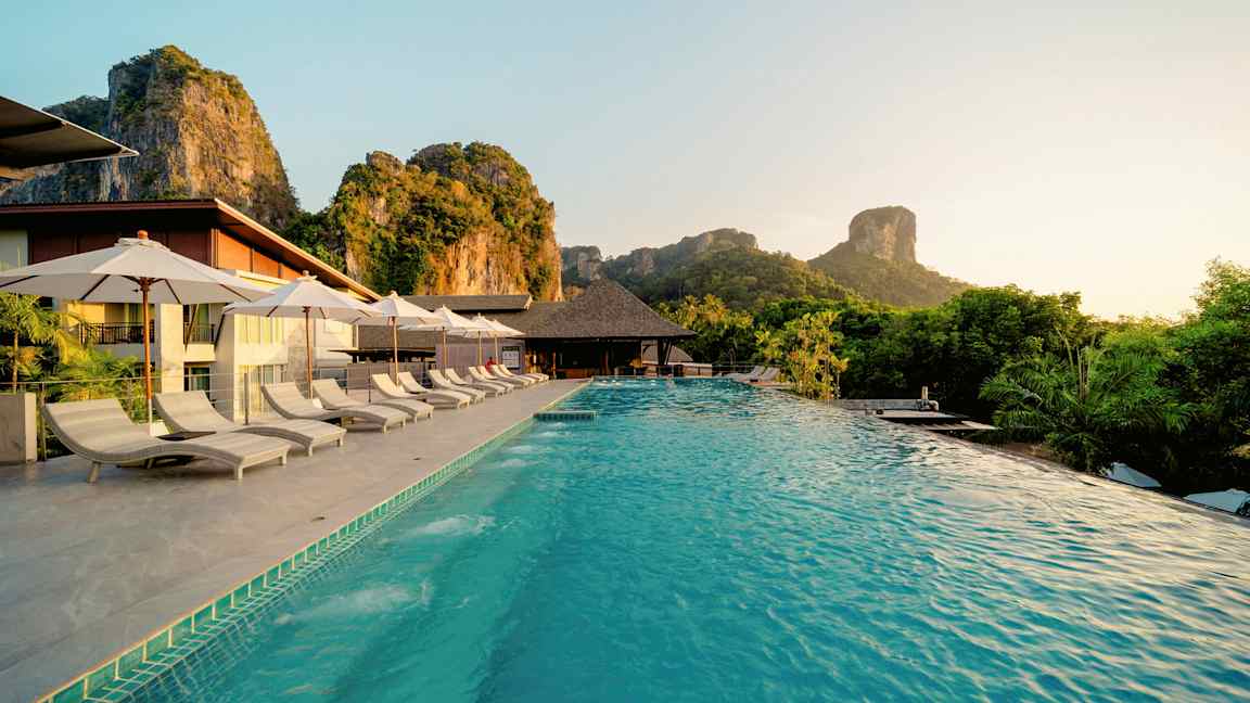 En pool med solstolar och parasoller vid en resort belägen i en naturskön miljö med berg i bakgrunden. (AI-genererad text)