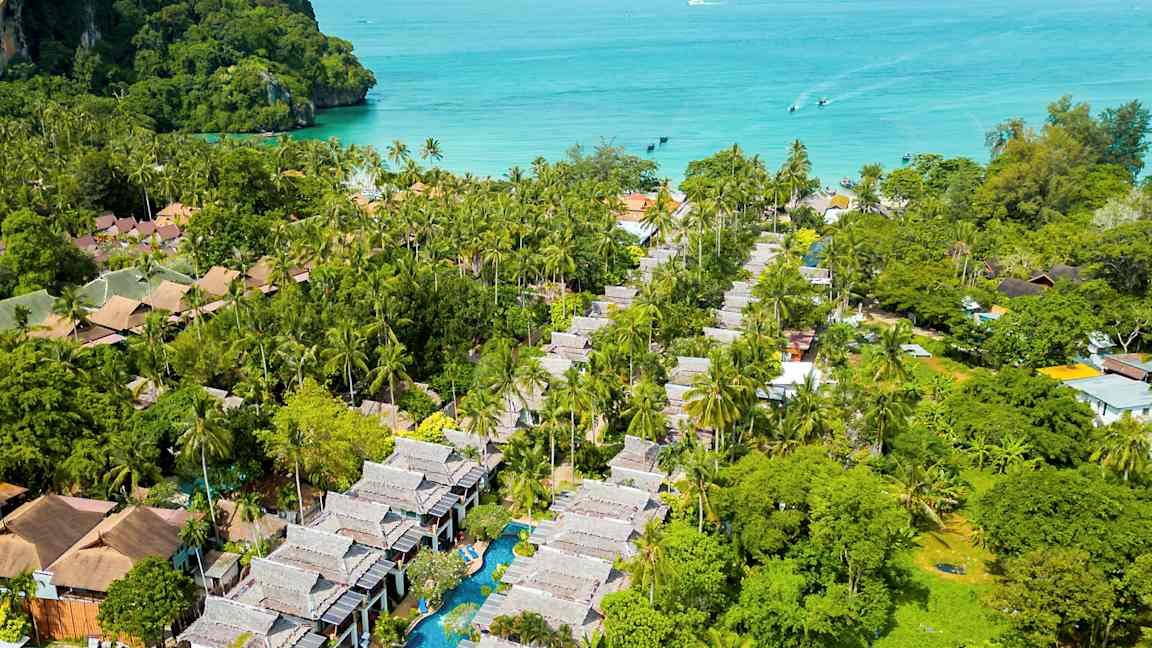 Flygbild över ett tropiskt område med palmer, byggnader och en pool, med havet i bakgrunden. (AI-genererad text)